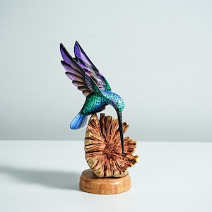 May include: A hand-carved wooden hummingbird sculpture featuring iridescent blue, green, and purple wings and body. The bird is positioned with its long, thin beak touching a textured, light brown wooden flower base set on a circular stand.