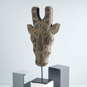 May include: A wooden giraffe head sculpture with a geometric pattern in black and gold. The sculpture is mounted on a black metal stand and a white base.