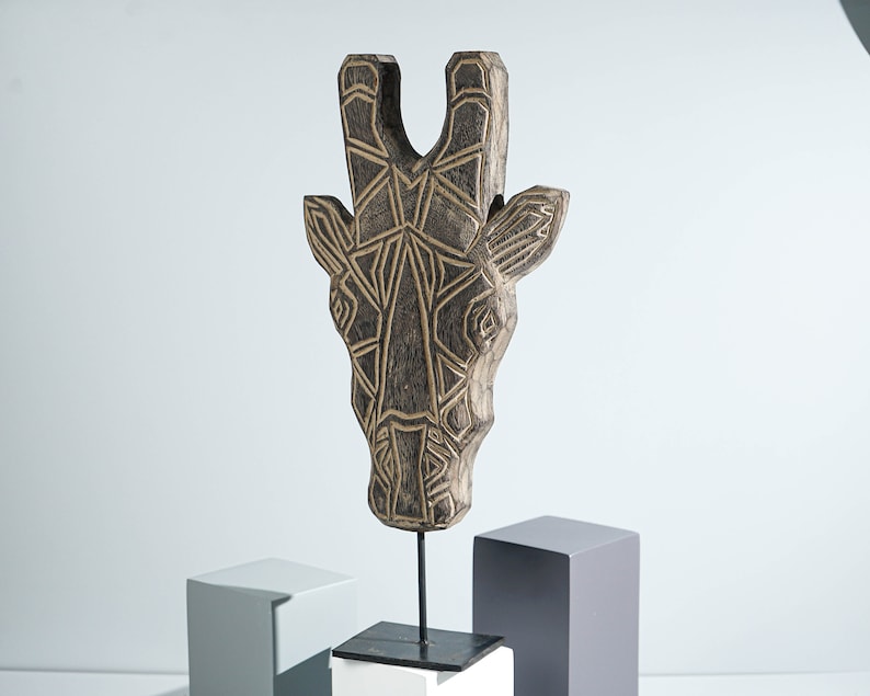May include: A wooden giraffe head sculpture with a geometric pattern in black and gold. The sculpture is mounted on a black metal stand and a white base.