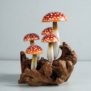 May include: A cluster of five decorative mushrooms with red caps and white spots. The mushrooms have white stems and are set in a piece of natural, dark brown wood. The background is a solid, light gray.