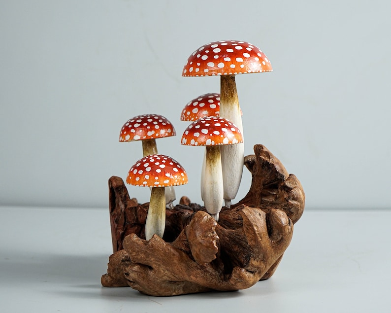 May include: A cluster of five decorative mushrooms with red caps and white spots. The mushrooms have white stems and are set in a piece of natural, dark brown wood. The background is a solid, light gray.
