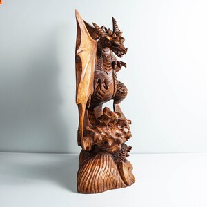 May include: A hand-carved wooden dragon sculpture featuring intricate scales, large wings, and a horned head. The dragon stands atop a textured, rocky base with detailed carvings. The wood displays a rich, polished brown grain throughout the entire piece.