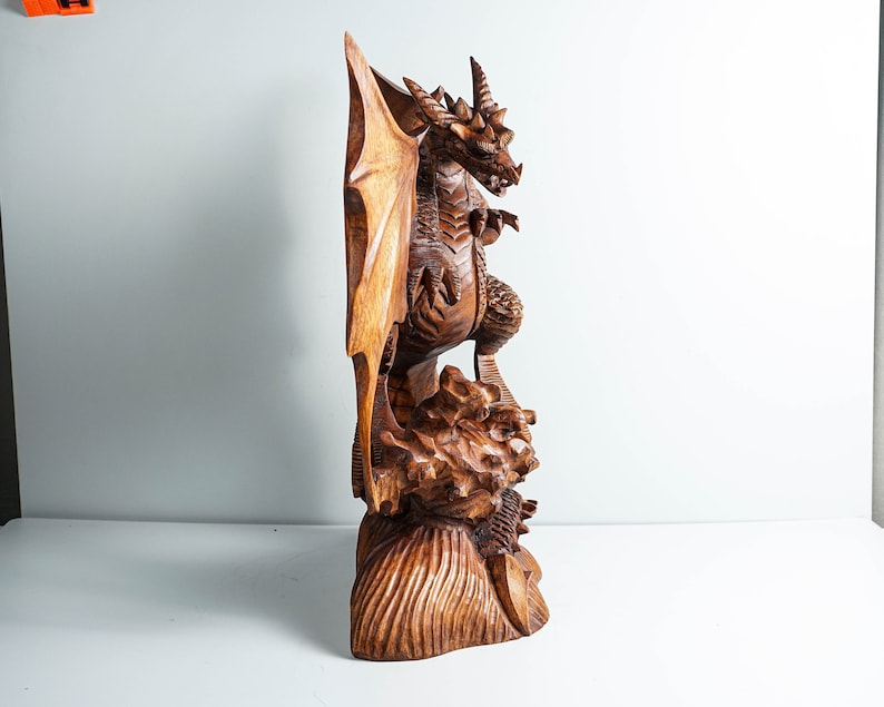 May include: A hand-carved wooden dragon sculpture featuring intricate scales, large wings, and a horned head. The dragon stands atop a textured, rocky base with detailed carvings. The wood displays a rich, polished brown grain throughout the entire piece.