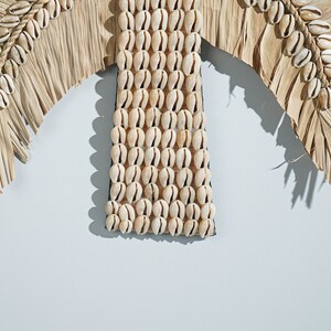 May include: A wall decor piece made of natural materials. It features a palm tree shape with brown woven leaves and a center section made of white seashells.