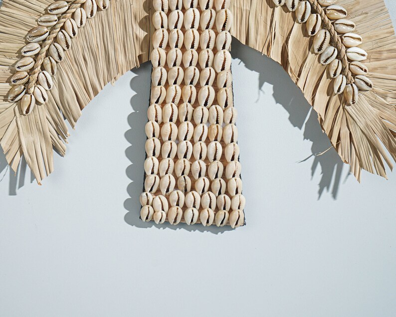 May include: A wall decor piece made of natural materials. It features a palm tree shape with brown woven leaves and a center section made of white seashells.