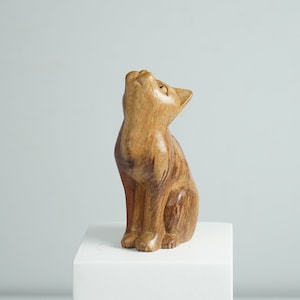 May include: A hand-carved wooden figurine of a cat in a sitting position with its head tilted upward. The sculpture features a natural light brown wood grain with visible texture, smooth contours, and a polished finish, capturing a feline form.