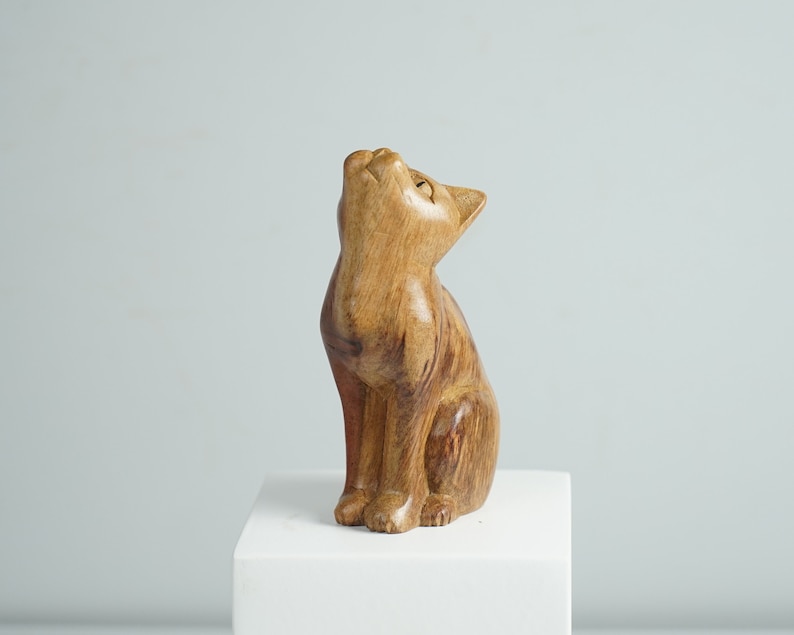 May include: A hand-carved wooden figurine of a cat in a sitting position with its head tilted upward. The sculpture features a natural light brown wood grain with visible texture, smooth contours, and a polished finish, capturing a feline form.