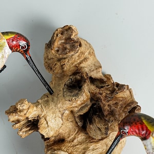 Puede incluir: Primer plano de una escultura de madera que presenta tres colibr&iacute;es con plumaje rojo, amarillo y verde. Las aves tienen largos picos negros y est&aacute;n posadas cerca de un trozo de madera texturizada de color marr&oacute;n claro.