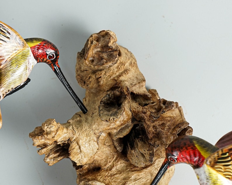 Puede incluir: Primer plano de una escultura de madera que presenta tres colibr&iacute;es con plumaje rojo, amarillo y verde. Las aves tienen largos picos negros y est&aacute;n posadas cerca de un trozo de madera texturizada de color marr&oacute;n claro.