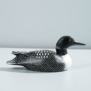 May include: A hand-carved wooden loon decoy featuring a solid black head with a small red eye, a white neck patch, and a black body covered in white polka dots and linear markings along the wings and tail feathers, presented in a side profile view.