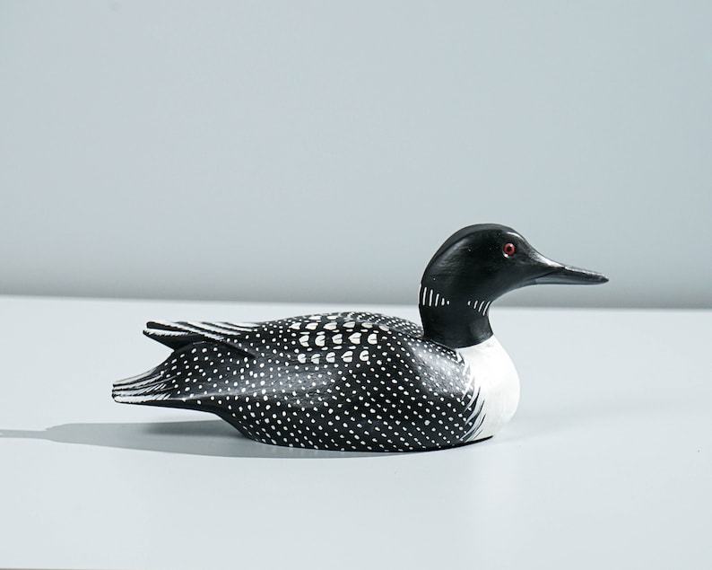 May include: A hand-carved wooden loon decoy featuring a solid black head with a small red eye, a white neck patch, and a black body covered in white polka dots and linear markings along the wings and tail feathers, presented in a side profile view.