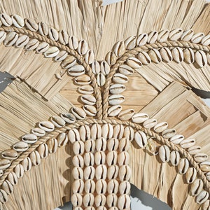 May include: A wall hanging made of woven palm leaves and cowrie shells. The shells are arranged in a pattern that resembles a palm tree.