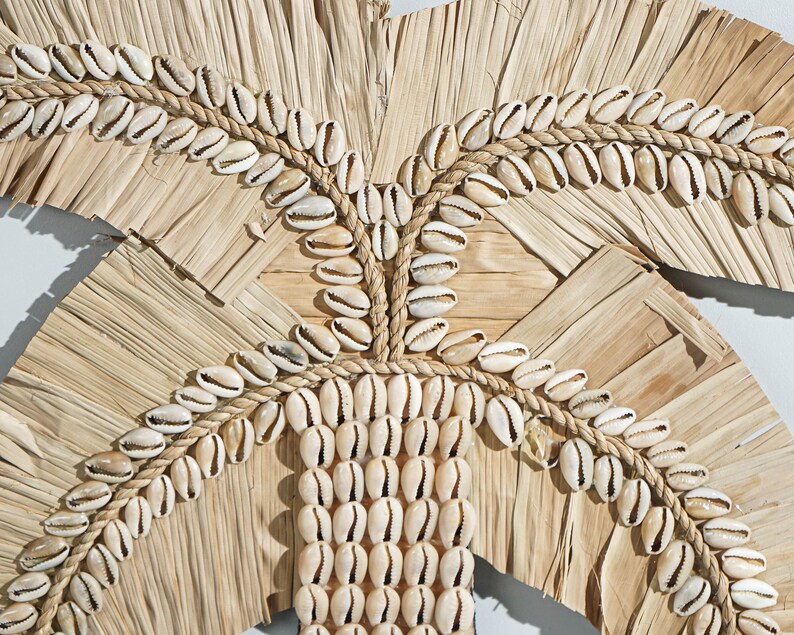 May include: A wall hanging made of woven palm leaves and cowrie shells. The shells are arranged in a pattern that resembles a palm tree.