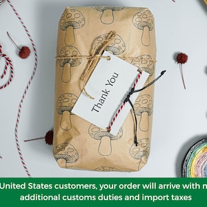 May include: A rectangular package wrapped in brown paper with a repeating mushroom pattern. A white tag with "Thank You" is attached with twine. Decorative elements include red and white twine and small, dried flower accents.