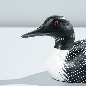 May include: A hand-carved wooden loon figurine featuring a black head, a sharp pointed beak, and a bright red eye. The neck has white vertical stripes, and the body is painted black with white spots across the back and wings, with a white chest.