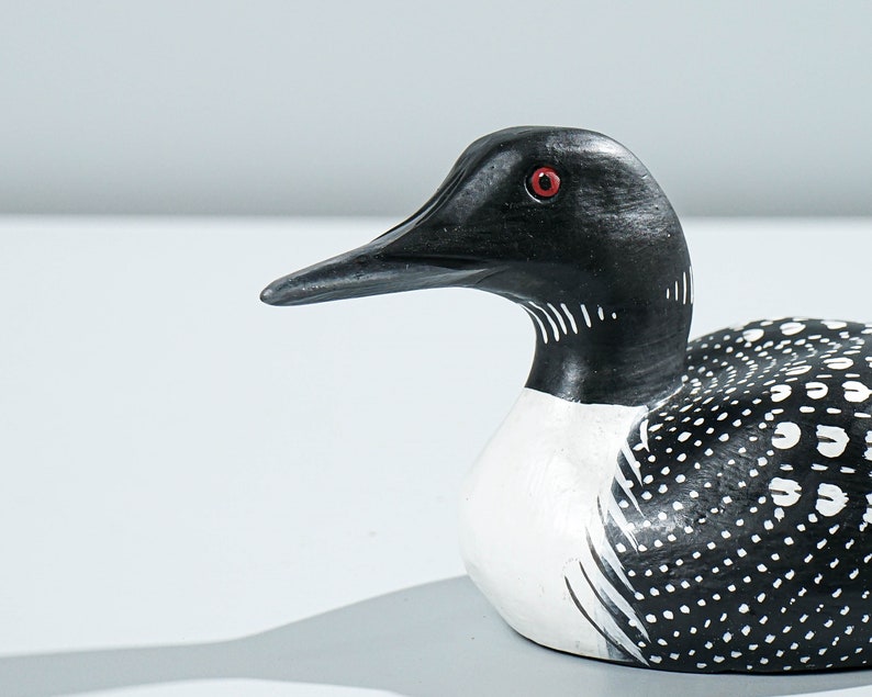 May include: A hand-carved wooden loon figurine featuring a black head, a sharp pointed beak, and a bright red eye. The neck has white vertical stripes, and the body is painted black with white spots across the back and wings, with a white chest.
