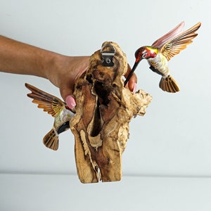 Puede incluir: Adorno de pared de madera con dos colibr&iacute;es tallados. Las aves tienen detalles rojos, amarillos y marrones. Se adjuntan a un trozo de madera natural con un colgador de metal.