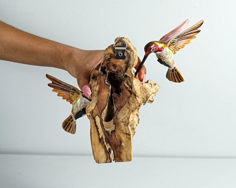 Puede incluir: Adorno de pared de madera con dos colibr&iacute;es tallados. Las aves tienen detalles rojos, amarillos y marrones. Se adjuntan a un trozo de madera natural con un colgador de metal.