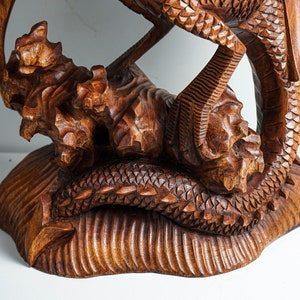 May include: A brown wooden sculpture featuring a detailed dragon with textured scales and sharp claws, perched upon a carved base that mimics a rocky or natural terrain. The wood grain is visible throughout the intricate, hand-carved surface.