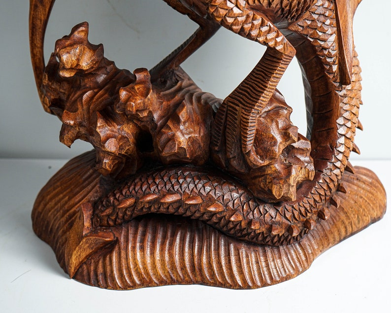 May include: A brown wooden sculpture featuring a detailed dragon with textured scales and sharp claws, perched upon a carved base that mimics a rocky or natural terrain. The wood grain is visible throughout the intricate, hand-carved surface.