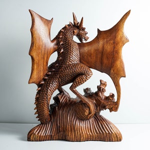 May include: A hand-carved wooden dragon sculpture featuring intricate scale detailing, large outstretched wings, and a spiked tail. The creature stands atop a textured, rocky base, with its head turned to the side, showcasing sharp horns and teeth.