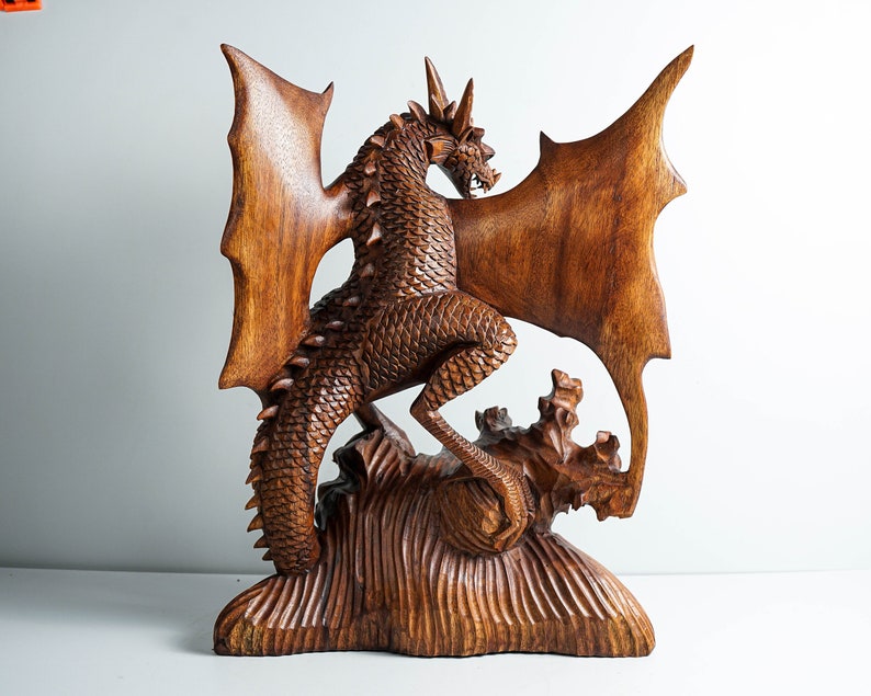 May include: A hand-carved wooden dragon sculpture featuring intricate scale detailing, large outstretched wings, and a spiked tail. The creature stands atop a textured, rocky base, with its head turned to the side, showcasing sharp horns and teeth.