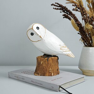 K&ouml;nnte beinhalten: Eine wei&szlig;e Holz-Eulenfigur mit schwarzen Augen und goldenen Details, die auf einem kleinen Holzstumpf sitzt. Die Eule steht auf einem Buch mit dem Text "#dear tomorrow" und "maudy ayunda".
