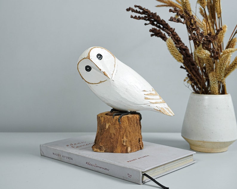 K&ouml;nnte beinhalten: Eine wei&szlig;e Holz-Eulenfigur mit schwarzen Augen und goldenen Details, die auf einem kleinen Holzstumpf sitzt. Die Eule steht auf einem Buch mit dem Text "#dear tomorrow" und "maudy ayunda".