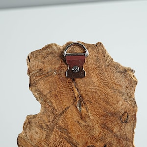 Puede incluir: Primer plano de la parte posterior de un colgante de pared de madera r&uacute;stica. La madera tiene un patr&oacute;n de vetas natural y texturizado con diferentes tonos de marr&oacute;n. Se adjunta una anilla en D y un soporte met&aacute;lico para colgar. El soporte es de color marr&oacute;n rojizo.