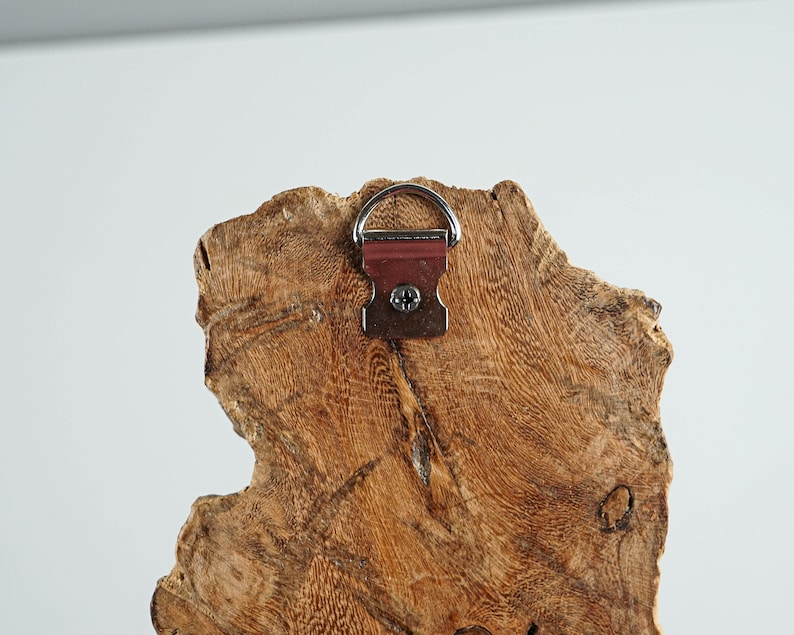 Puede incluir: Primer plano de la parte posterior de un colgante de pared de madera r&uacute;stica. La madera tiene un patr&oacute;n de vetas natural y texturizado con diferentes tonos de marr&oacute;n. Se adjunta una anilla en D y un soporte met&aacute;lico para colgar. El soporte es de color marr&oacute;n rojizo.