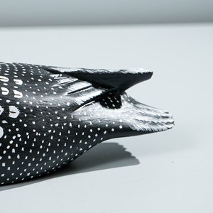May include: A carved wooden bird figurine featuring a black body with white painted spots and markings. The tail section shows detailed carved feather textures with white highlights along the edges, contrasting against the dark black base color.