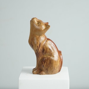 May include: A hand-carved wooden cat figurine with a smooth, polished finish and visible natural wood grain patterns. The cat is depicted in a seated position with its head tilted upward, featuring pointed ears and carved facial details.
