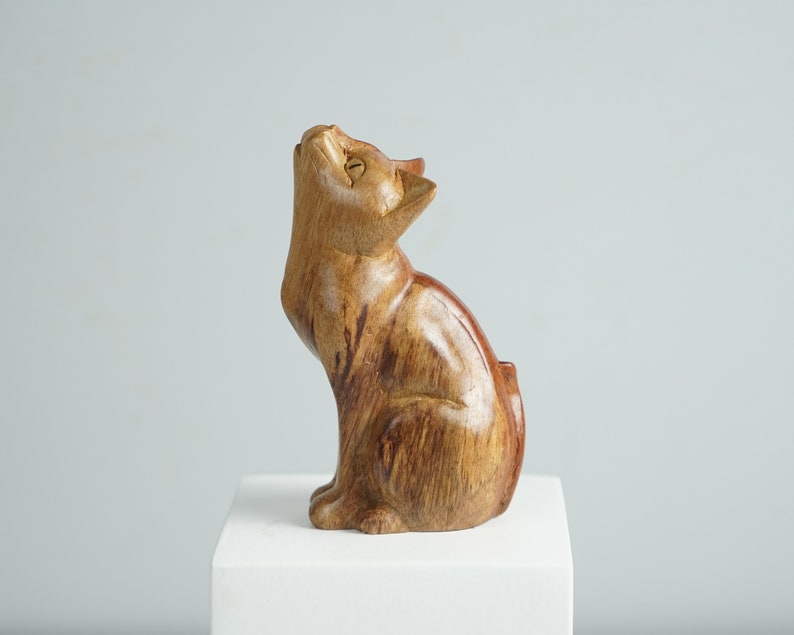 May include: A hand-carved wooden cat figurine with a smooth, polished finish and visible natural wood grain patterns. The cat is depicted in a seated position with its head tilted upward, featuring pointed ears and carved facial details.