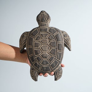 Könnte beinhalten: Eine dunkelbraune Holzschildkröte mit eingravierten, hellen geometrischen Mustern auf dem Panzer, dem Kopf und den Flossen. Die Holzoberfläche weist eine sichtbare Maserung auf, und die geschnitzten Linien bilden einen deutlichen Kontrast zum dunklen Finish der Schildkrötenfigur.