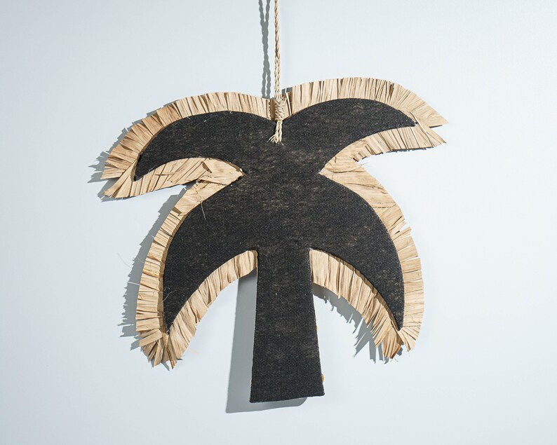 May include: A black palm tree cutout with a fringed raffia border. The palm tree is hanging from a string.