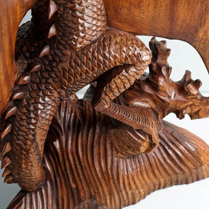 May include: A hand-carved wooden sculpture of a dragon with detailed scales and spikes along its back. The dragon is depicted in a dynamic pose with its tail curled and wings extended, resting on a textured base carved from the same dark brown wood.