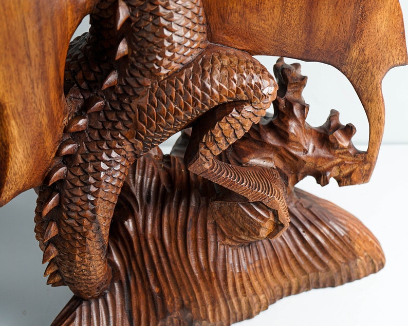 May include: A hand-carved wooden sculpture of a dragon with detailed scales and spikes along its back. The dragon is depicted in a dynamic pose with its tail curled and wings extended, resting on a textured base carved from the same dark brown wood.