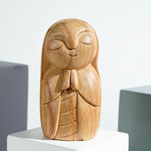 May include: A light brown wooden figurine carved into a smooth, rounded shape with a serene facial expression, closed eyes, and hands pressed together in a prayer position. The wood grain is visible throughout the surface of the carved sculpture.
