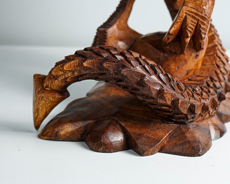 May include: A wooden sculpture of a dragon with intricate details. The dragon is carved in a dark brown color and has a textured surface. The dragon's tail is curled around its body, and its head is raised up.