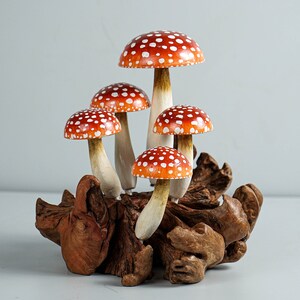 May include: A cluster of five decorative mushrooms with red caps and white spots. The mushrooms have beige stems and are attached to a piece of natural wood. The caps are a vibrant red-orange color.