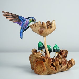 May include: A hand-carved wooden sculpture featuring a vibrant blue and purple hummingbird hovering over a large wooden flower. The base is a piece of textured driftwood with three small green mushrooms with white spots growing from it.