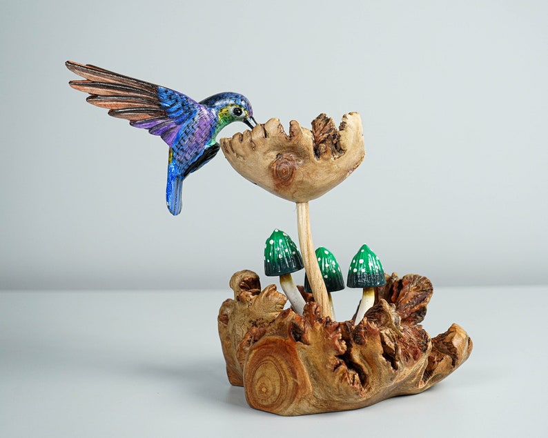 May include: A hand-carved wooden sculpture featuring a vibrant blue and purple hummingbird hovering over a large wooden flower. The base is a piece of textured driftwood with three small green mushrooms with white spots growing from it.