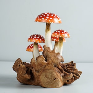 May include: A cluster of wooden mushrooms with red caps and white spots, mounted on a piece of natural wood. The mushroom stems are white with a brown base. The wood base has a natural, organic shape.