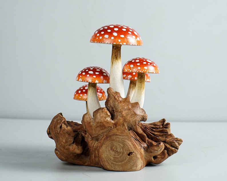 May include: A cluster of wooden mushrooms with red caps and white spots, mounted on a piece of natural wood. The mushroom stems are white with a brown base. The wood base has a natural, organic shape.