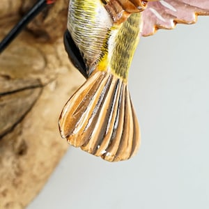 Puede incluir: Primer plano de un p&aacute;jaro de madera tallada. El p&aacute;jaro tiene un cuerpo amarillo y verde, con alas marrones y blancas. Las plumas de la cola son marrones y blancas. El p&aacute;jaro est&aacute; montado sobre un trozo de madera natural.