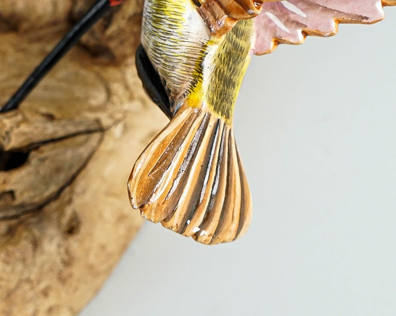 Puede incluir: Primer plano de un p&aacute;jaro de madera tallada. El p&aacute;jaro tiene un cuerpo amarillo y verde, con alas marrones y blancas. Las plumas de la cola son marrones y blancas. El p&aacute;jaro est&aacute; montado sobre un trozo de madera natural.