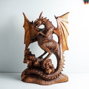 May include: A hand-carved wooden dragon sculpture featuring intricate scale detailing, large spread wings, and a long tail coiled around a rocky base. The natural brown wood grain is visible throughout the entire piece, highlighting the textured surface.