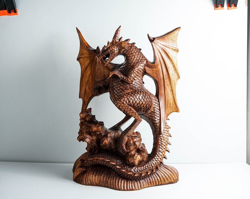 May include: A hand-carved wooden dragon sculpture featuring intricate scale detailing, large spread wings, and a long tail coiled around a rocky base. The natural brown wood grain is visible throughout the entire piece, highlighting the textured surface.