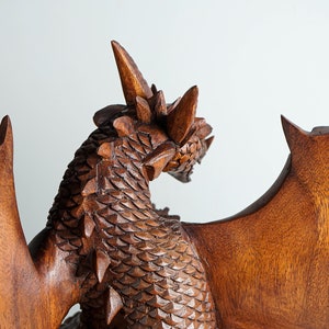 May include: A brown wooden dragon sculpture featuring intricate carved scales along the neck and back, prominent horns on the head, and large, smooth, outstretched wings. The wood grain is visible throughout the detailed, textured surface.