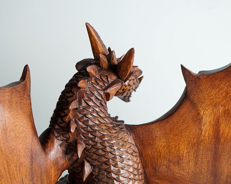May include: A brown wooden dragon sculpture featuring intricate carved scales along the neck and back, prominent horns on the head, and large, smooth, outstretched wings. The wood grain is visible throughout the detailed, textured surface.